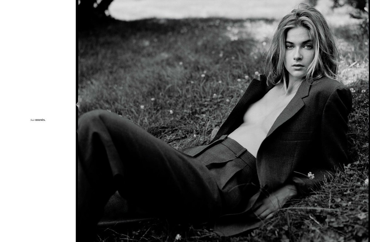 Black-and-white fashion shot of a young woman in a blazer and trousers lying in the grass and looking at the camera.