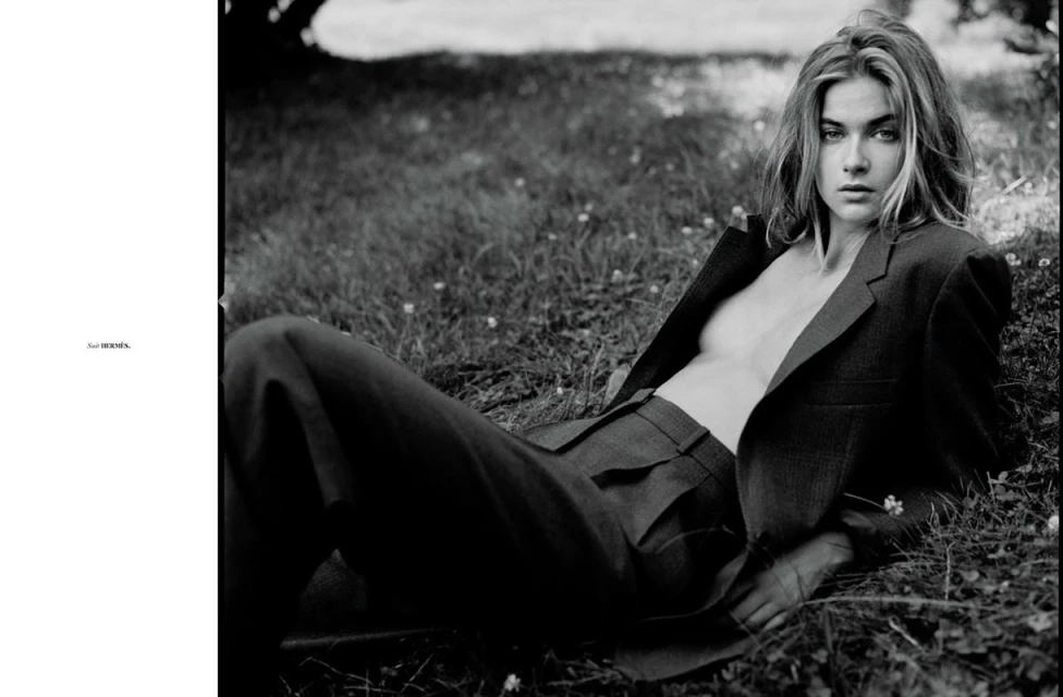 Black-and-white fashion shot of a young woman in a blazer and trousers lying in the grass and looking at the camera.