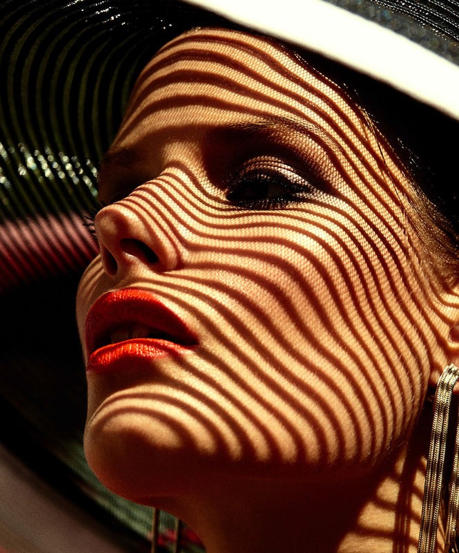 Close-up of woman's face with red lips and black hat, featuring shadow pattern from hat. Beauty shot with artistic lighting.