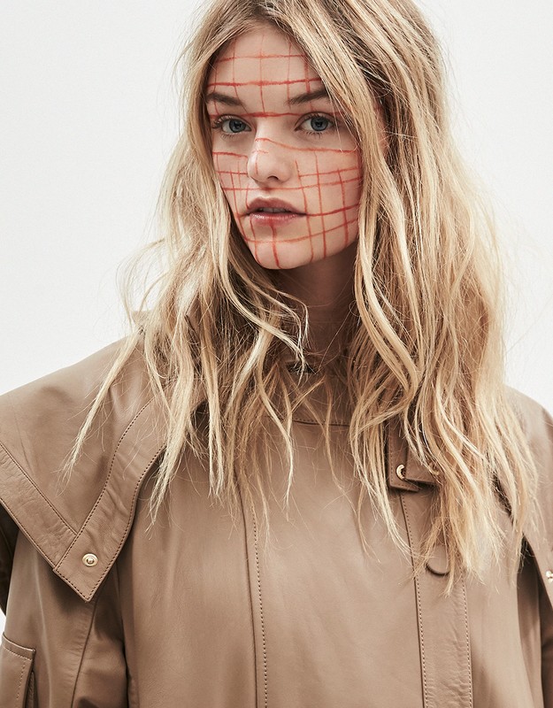 Close-up shot of a blonde adult female with a red grid design on her face, wearing a tan jacket in a brightly lit photo studio.