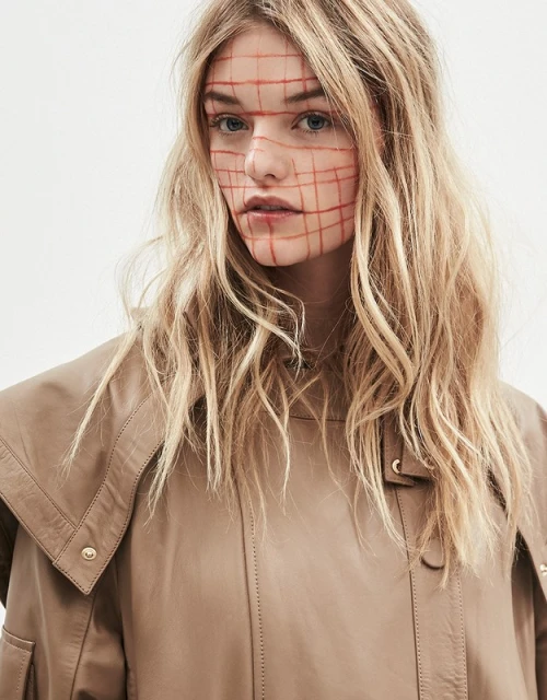 Close-up shot of a blonde adult female with a red grid design on her face, wearing a tan jacket in a brightly lit photo studio.