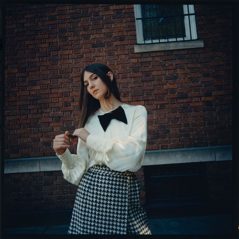 Young woman in a cream blouse with a bow and houndstooth skirt stands outdoors against a brick wall on a city street.
