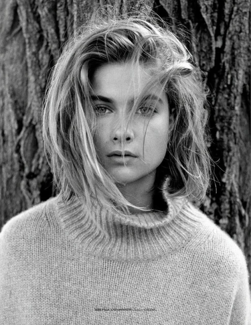 Black and white portrait of a young woman with short blonde hair and a turtleneck sweater posing in front of a textured tree.