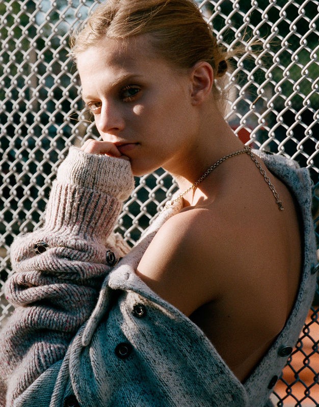 Portrait of a blond female adult with her hand to her face, wearing a cardigan, against a chain-link fence outdoors on a sunny day.