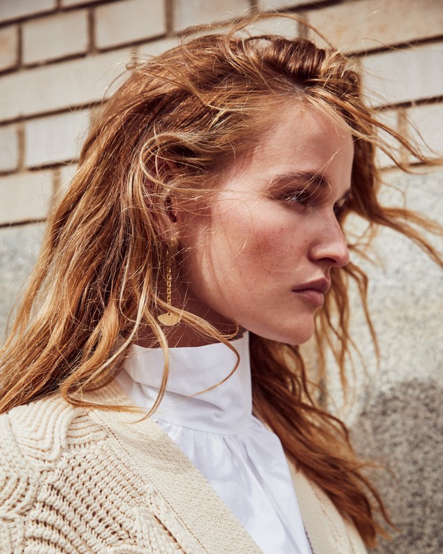 Close up portrait of a woman with long windblown blonde hair wearing a white high neck top and textured cream cardigan