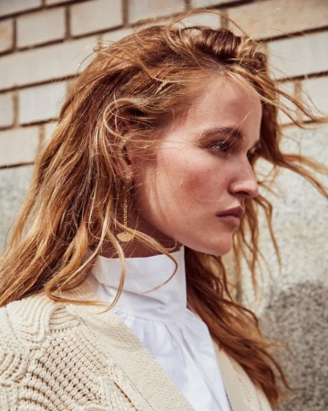 Close up portrait of a woman with long windblown blonde hair wearing a white high neck top and textured cream cardigan
