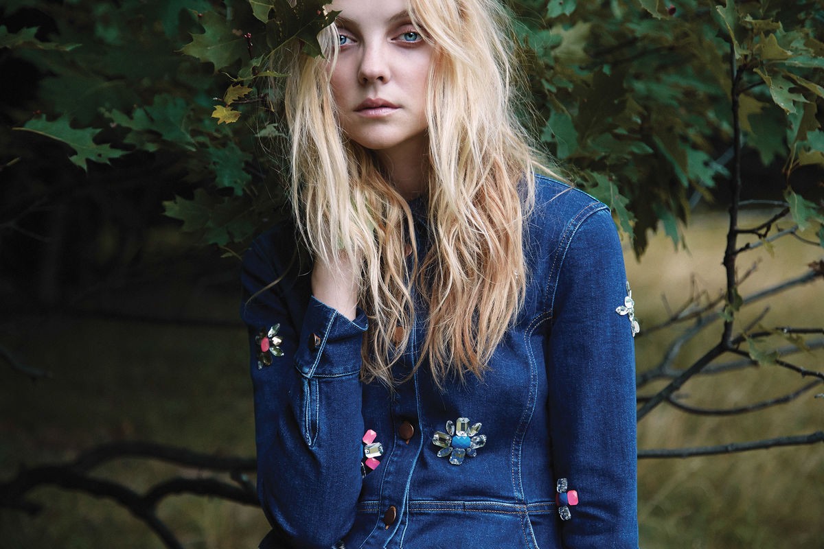 Blonde woman in a denim jacket poses in front of lush green foliage, her hand near her face, gazing slightly off camera.
