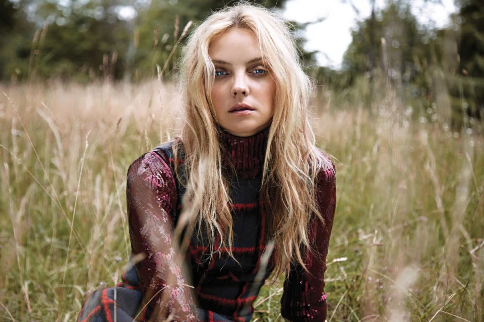 A woman with blonde hair sits in a grassy field, wearing a plaid dress and sequined top, photographed in a natural outdoor setting.