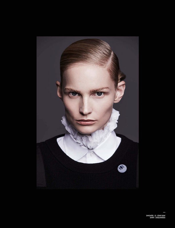 Headshot of a person with slicked back blonde hair wearing a white ruffled collar and black sweater against a grey background.
