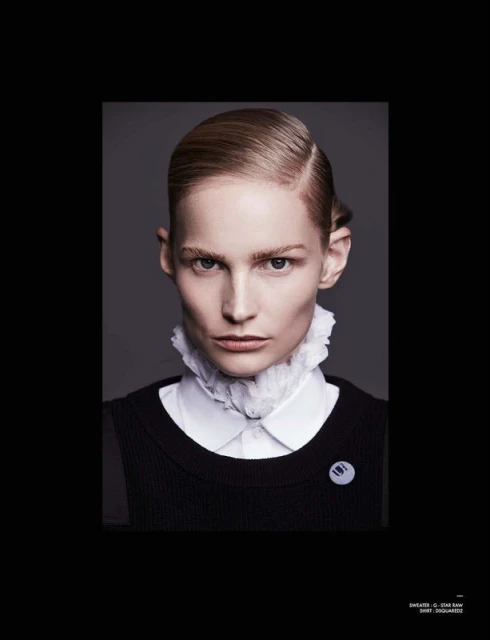Headshot of a person with slicked back blonde hair wearing a white ruffled collar and black sweater against a grey background.