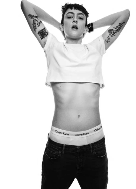 Black and white portrait of a person with tattoos wearing a crop top and jeans with hands behind the head in a studio setting.
