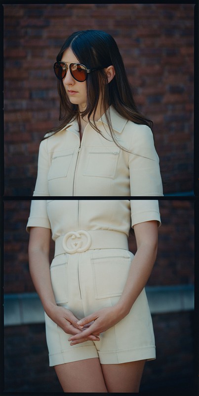 Brunette woman in sunglasses and off-white romper with belt poses against a brick wall backdrop for a fashion editorial.