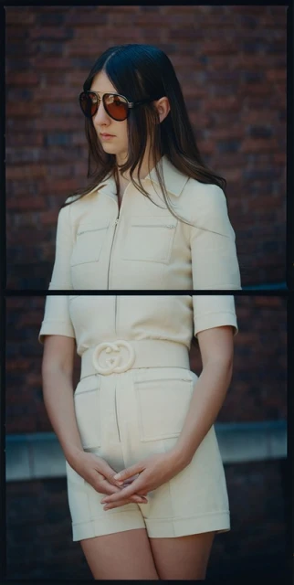 Brunette woman in sunglasses and off-white romper with belt poses against a brick wall backdrop for a fashion editorial.