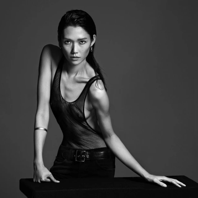Monochrome portrait of an Asian woman in a mesh top and jeans, looking directly at the camera against a plain gray background.