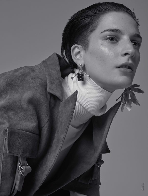 A young adult woman with short, slicked-back hair wearing a suede jacket and turtleneck, accessorized with artistic earrings, in studio lighting.