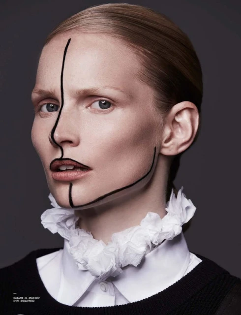 Portrait of a fair-skinned woman with a white ruffled collar and black lines painted on her face against a gray backdrop.