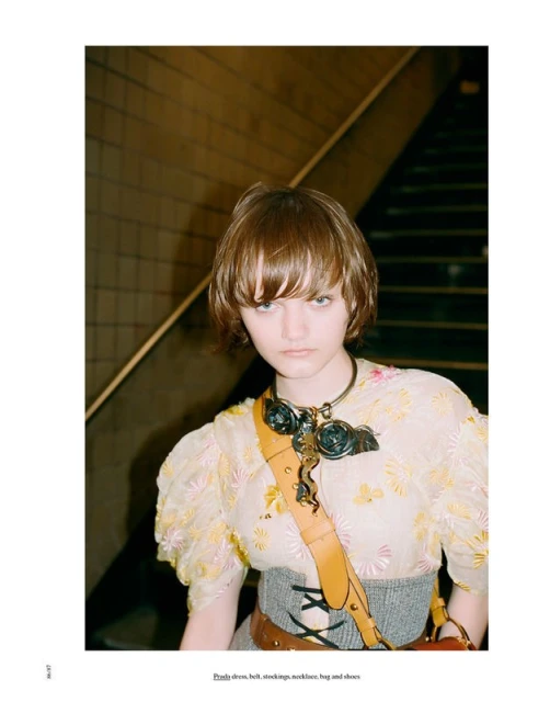 Indoor fashion portrait of a young adult female with short brown hair, wearing an elaborate steampunk-style necklace and floral dress.