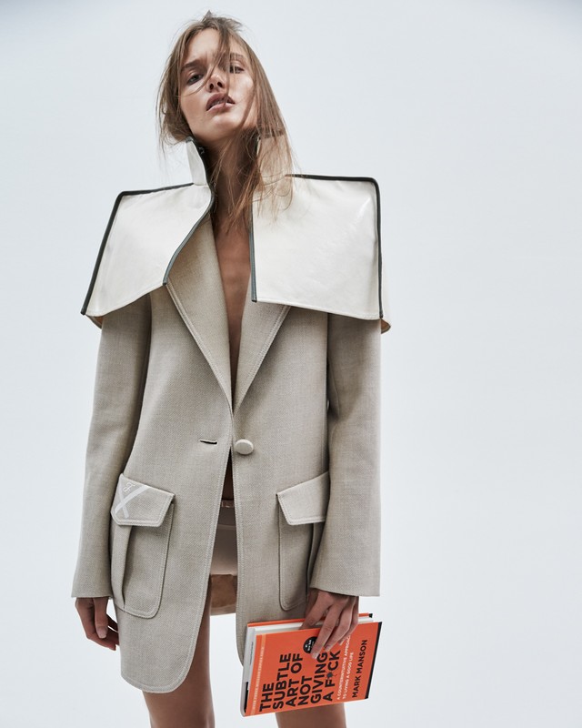 Fashion photo of a woman wearing an oversized beige blazer with a white collar detail, holding a book in a bright white photo studio.
