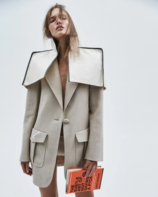 Fashion photo of a woman wearing an oversized beige blazer with a white collar detail, holding a book in a bright white photo studio.