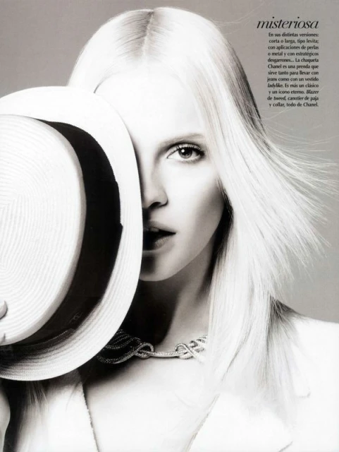 Monochrome portrait of a model with platinum blonde hair, wearing a Chanel necklace, partially obscured by a wide-brimmed hat.