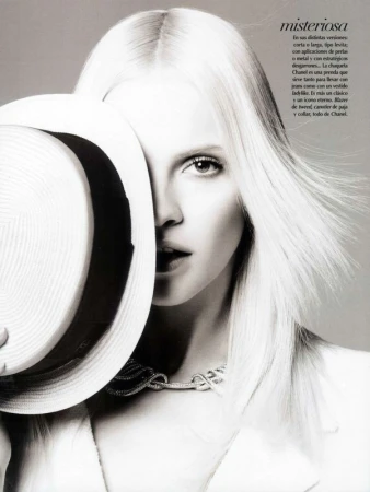Monochrome portrait of a model with platinum blonde hair, wearing a Chanel necklace, partially obscured by a wide-brimmed hat.