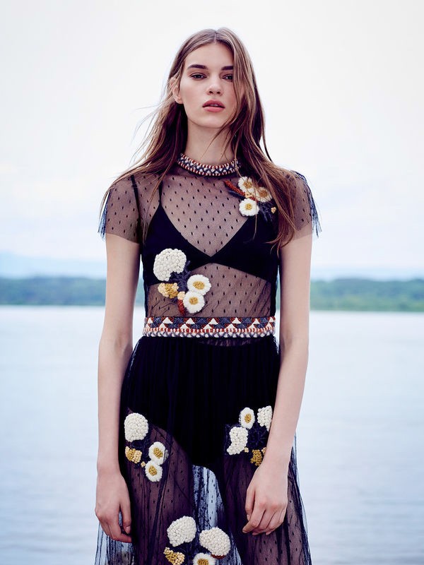 A fashion model poses in a sheer black dress with floral accents against a blurred background of water and trees in an outdoor setting.