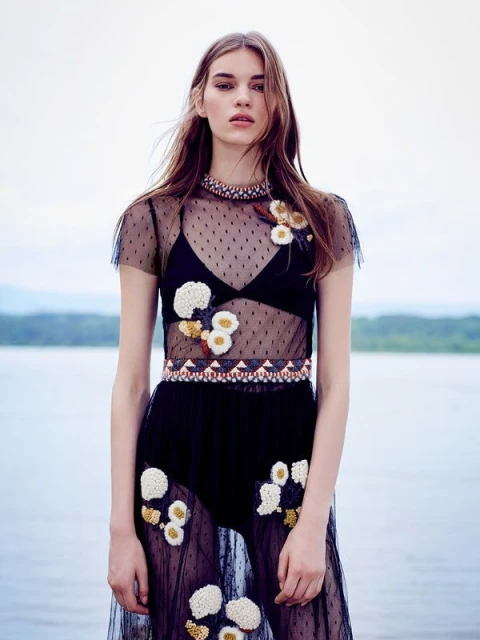 A fashion model poses in a sheer black dress with floral accents against a blurred background of water and trees in an outdoor setting.