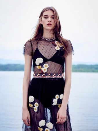 A fashion model poses in a sheer black dress with floral accents against a blurred background of water and trees in an outdoor setting.