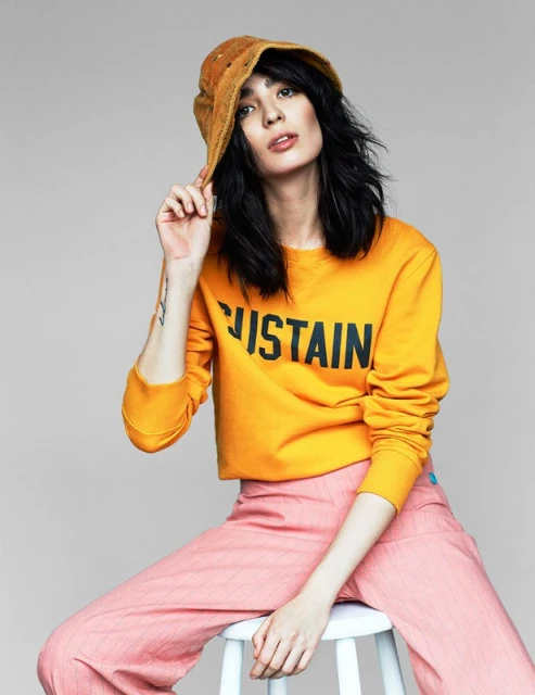 Young woman in a yellow shirt and pink pants sits on a white stool, wearing a hat in a studio setting. Fashion editorial shot.