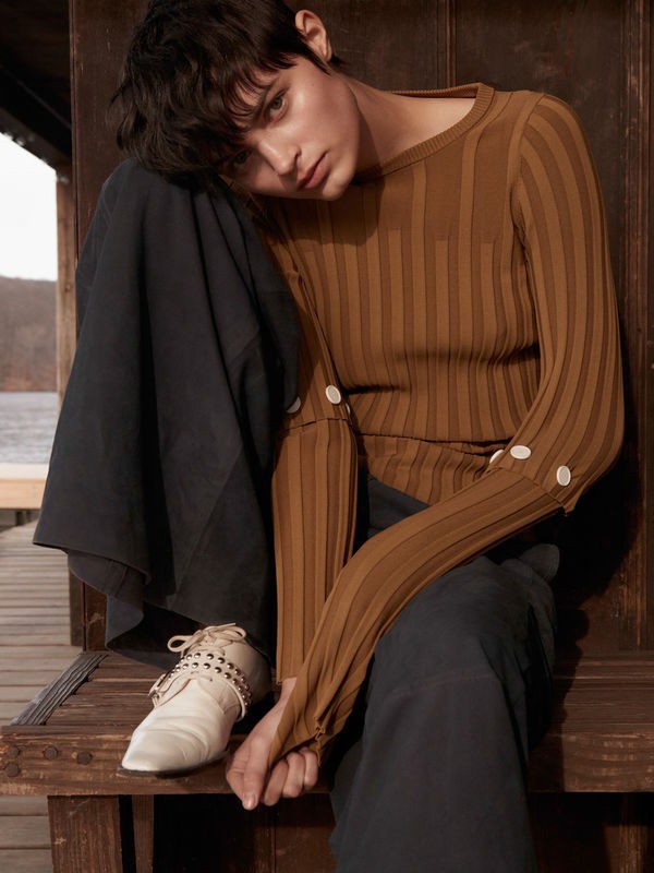 Fashion shot of a woman with short dark hair wearing a brown ribbed long-sleeved shirt and dark pants, sitting on wooden steps outdoors.