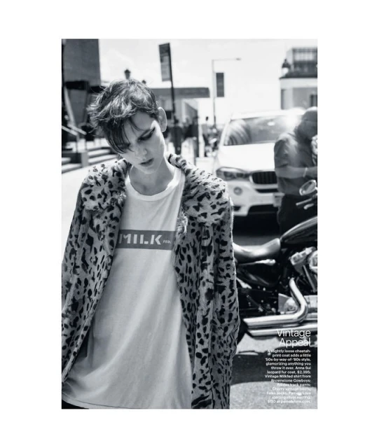 Monochrome fashion shot of a person in a white tee shirt and leopard print jacket standing next to a motorcycle on a city street.
