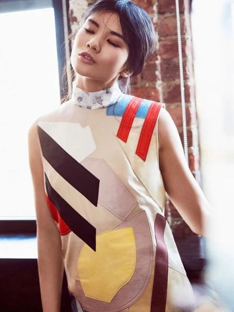 Portrait of an Asian female model in a sleeveless, patchwork-design top, posing indoors by a window with a brick wall in the background.