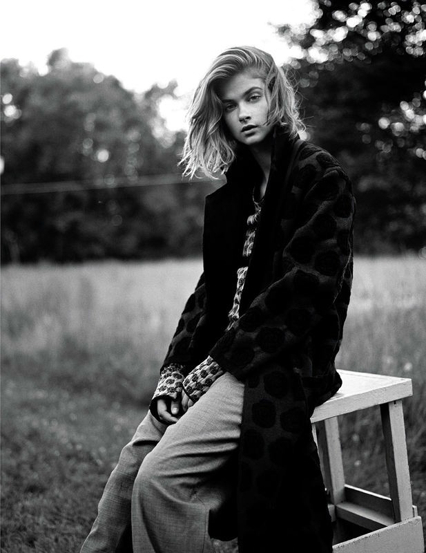 Monochrome portrait of a blonde woman in a patterned coat, sitting on a wooden stool outdoors in a blurred grassy field setting.