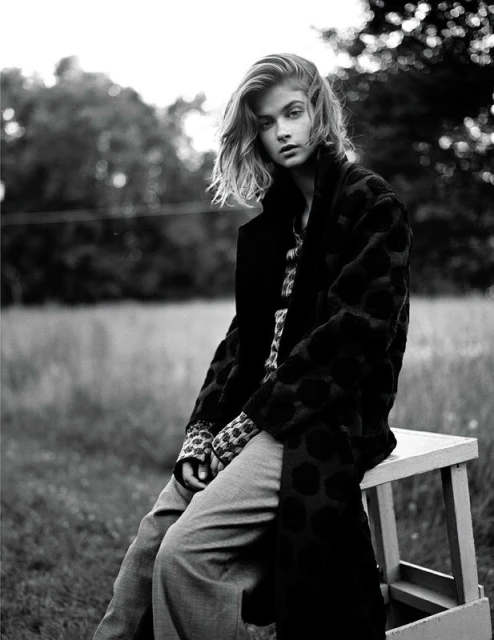 Monochrome portrait of a blonde woman in a patterned coat, sitting on a wooden stool outdoors in a blurred grassy field setting.