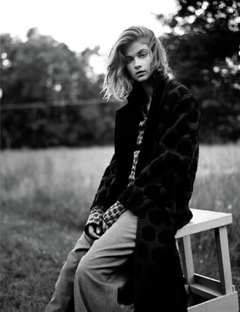 Monochrome portrait of a blonde woman in a patterned coat, sitting on a wooden stool outdoors in a blurred grassy field setting.