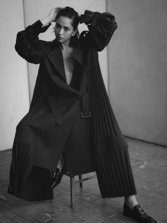 Monochrome fashion shot of a woman in a coat and wide-leg pants, seated on a stool, holding her hair, with a gray background.