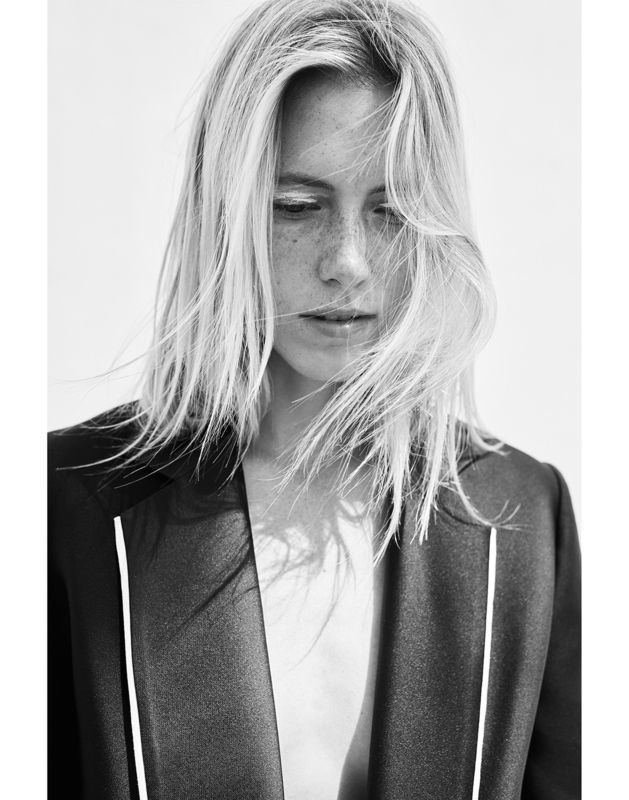 Monochrome portrait of a fair-skinned woman with blonde hair wearing a tailored black jacket in a minimalist studio setting.