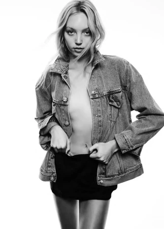 Black and white portrait of a female model wearing an oversized denim jacket over dark shorts against a bright background.