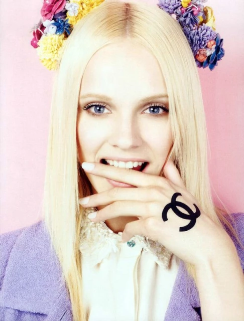 Close-up of a smiling blonde woman with flower headpiece and Chanel logo tattoo on hand against a light pink background.