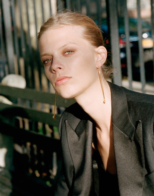 Portrait of a blonde woman with long earrings wearing a black blazer against an urban background with buildings and a green bench.