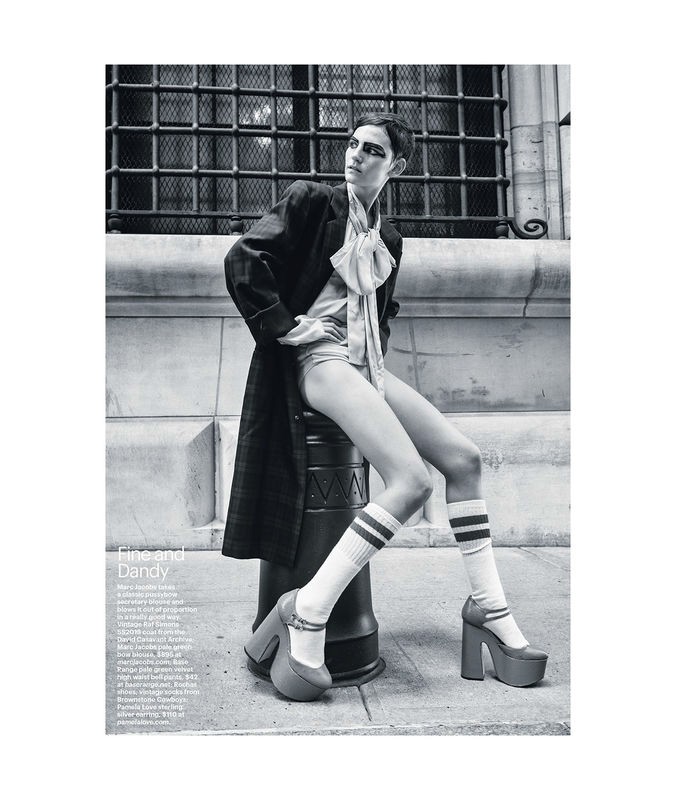 Black and white fashion photograph of a woman in a coat and shorts sitting on a metal bollard wearing platform shoes and knee-high socks.