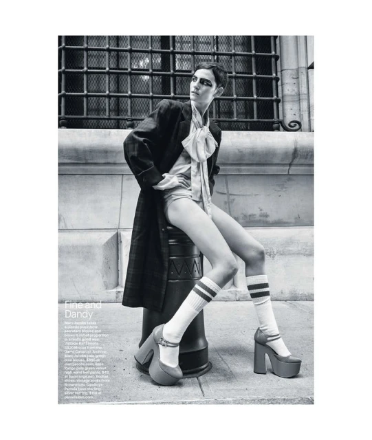 Black and white fashion photograph of a woman in a coat and shorts sitting on a metal bollard wearing platform shoes and knee-high socks.