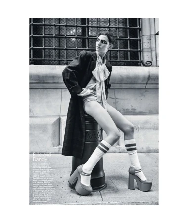 Black and white fashion photograph of a woman in a coat and shorts sitting on a metal bollard wearing platform shoes and knee-high socks.