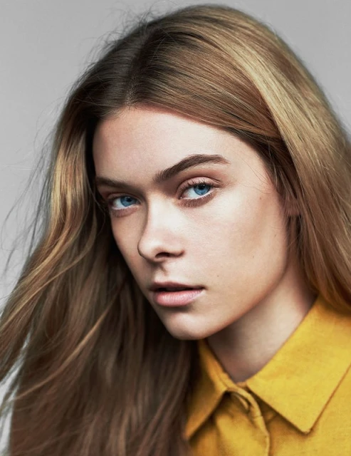 Portrait of a young woman with long blonde hair and blue eyes wearing a yellow collared shirt against a neutral background, close-up.