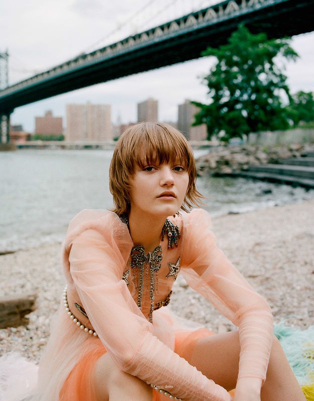 A young woman in a peach dress sits on a shore with a bridge and city buildings in the background on an overcast day. 