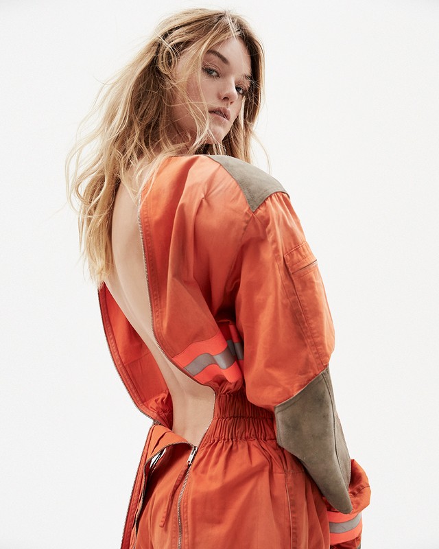 A woman in an orange jumpsuit looks back over her shoulder in a medium shot against a bright white background in a fashion editorial.