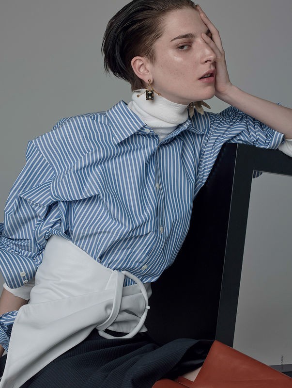 Fashion photo of a person with short dark hair, wearing a blue striped shirt and a white garment, sitting on a black chair in a studio.