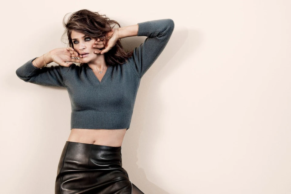 Brunette woman in a grey sweater and black leather skirt poses against a neutral wall with her hands near her face.