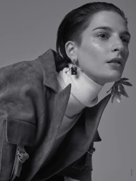 Close-up portrait of a woman with slicked-back hair wearing a suede jacket and large architectural earrings, set against a grey background.