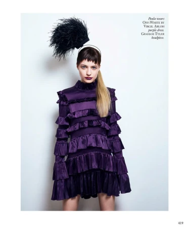 Model in ruffled purple dress and black feather headpiece against white seamless background